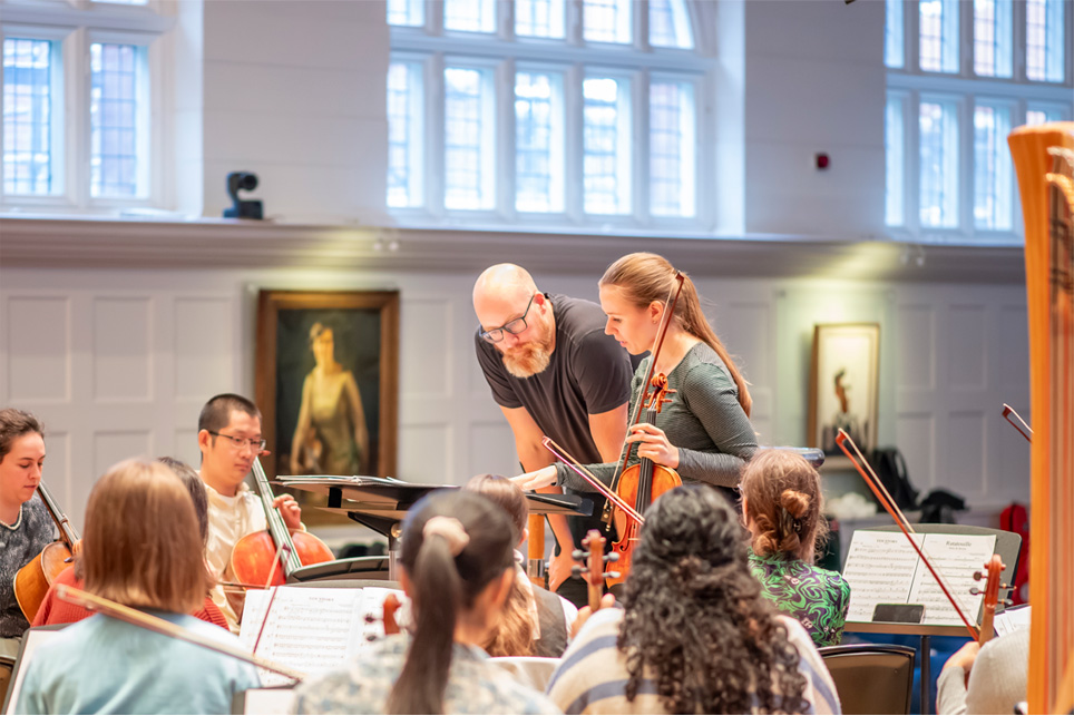 A female violinist standing next to a male conductor, looking at the conducting score, with the orchestra around then, on stage in a performance space.